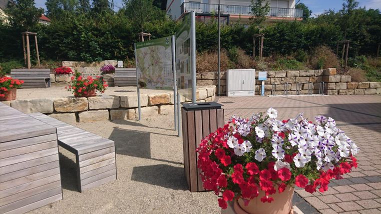 Een mooie buitenruimte met houten zitplaatsen en kleurrijke bloemen. Op de achtergrond staat een informatieschild en groene struiken omringen de sfeer.