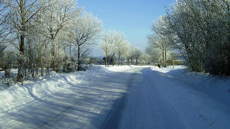Eine verschneite Straße, umgeben von frostigen Bäumen. Der Himmel ist klar und blau.