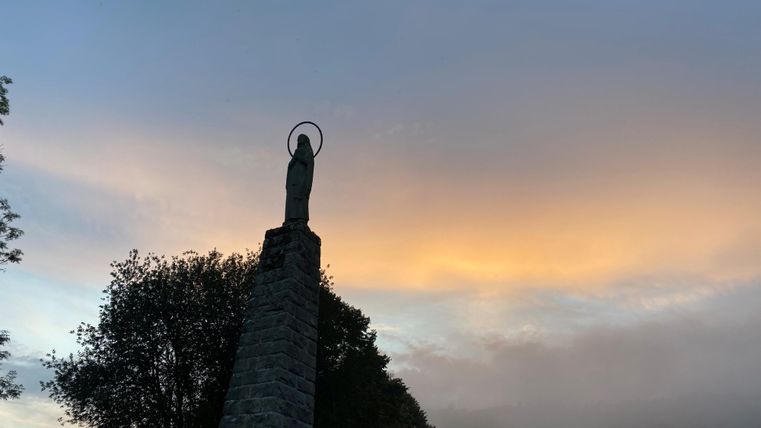 Eine Statue steht auf einem hohen Stein mit einem sanften, bunten Himmel im Hintergrund. Bäume umgeben die Statue und es ist leicht neblig.