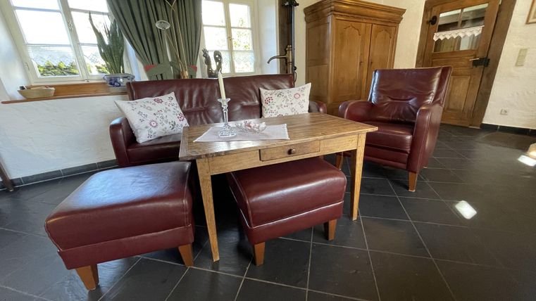 Un salon confortable avec un canapé en cuir rouge et deux fauteuils assortis. Sur la table en bois, il y a une nappe, et les fenêtres laissent entrer beaucoup de lumière.