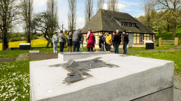 Eine Gruppe von Menschen versammelt sich vor einem historischen Gebäude in einer grünen Landschaft. Im Vordergrund ist ein graues Monument zu sehen.