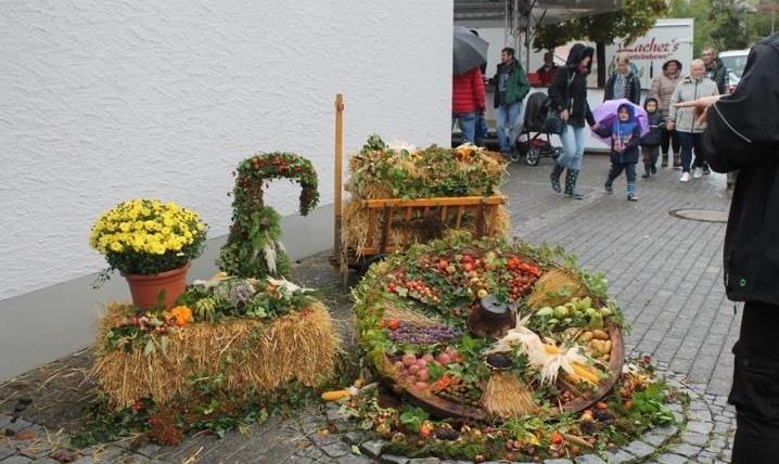 Ein bunter Erntedankaufsatz mit verschiedenen Gemüse- und Obstsorten, dekoriert mit Blumen und Gras. Im Hintergrund sind einige Menschen zu sehen, die die Szene beobachten.