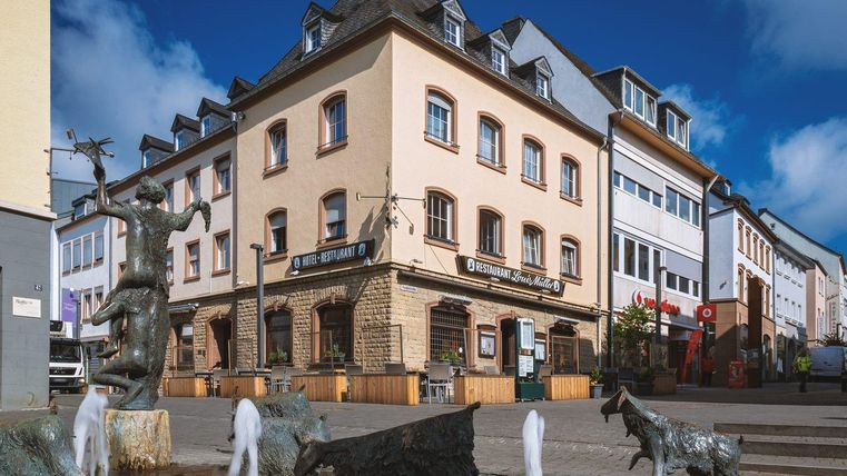 Eine hübsche Stadtansicht mit einem Gebäude im Vordergrund und einer Brunnenplastik. Die Straßen sind bevölkert und der Himmel ist klar und blau.