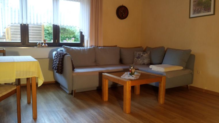 Un salon confortable avec un canapé gris et une table en bois. Les fenêtres laissent entrer beaucoup de lumière naturelle et il y a une atmosphère relaxante.