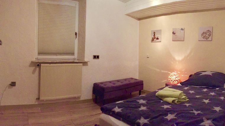 A cozy bedroom with a bed, a bench, and a window. Pictures hang on the wall and a soft light creates a pleasant atmosphere.