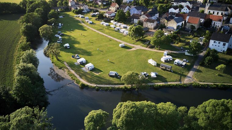 Een camping aan de rivier met veel campers en tenten. Op de achtergrond zijn huizen en een groene omgeving te zien.