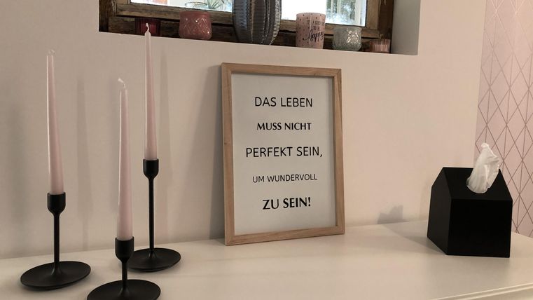 Ein stilvoller Tisch mit Kerzenhaltern und einem Rahmen mit dem Spruch: „Das Leben muss nicht perfekt sein, um wunderbar zu sein.“ Daneben befindet sich eine schwarze Box mit Taschentüchern.