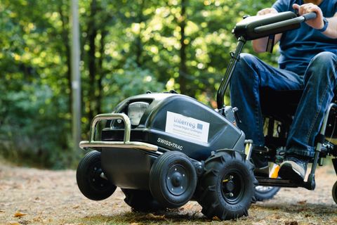 Ein motorisiertes Gerät für den Außenbereich, das an einem Rollstuhl befestigt ist. Es wird auf unebenem Boden im Wald verwendet.