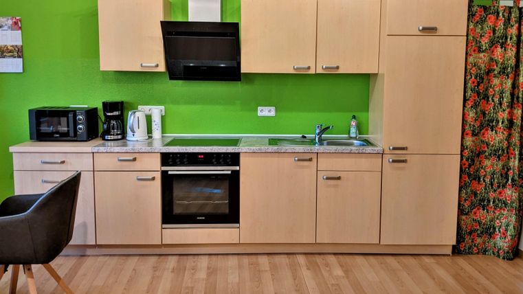 Eine moderne Küche mit Holzschränken und grüner Wand. Es gibt eine Mikrowelle, einen Wasserkocher und einen Herd.