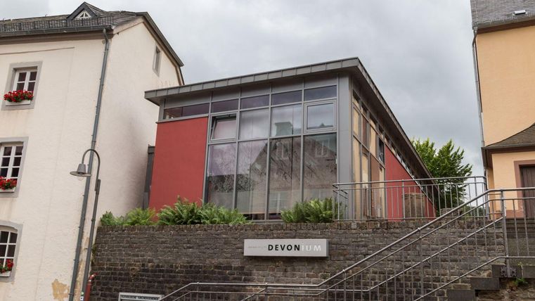 Ein modernes Gebäude mit großen Fenstern und einer roten Fassade. Es befindet sich an einer Treppe und ist von grünen Pflanzen umgeben.