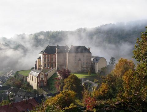 Ein malerisches Schloss umgeben von Nebel und Bäumen in herbstlichen Farben. Die sanfte Hügellandschaft trägt zur idyllischen Atmosphäre bei.