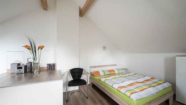 Une chambre moderne avec un lit double et une literie colorée. La pièce a des murs clairs et des détails en bois, ainsi qu'une chaise et un petit coin cuisine.
