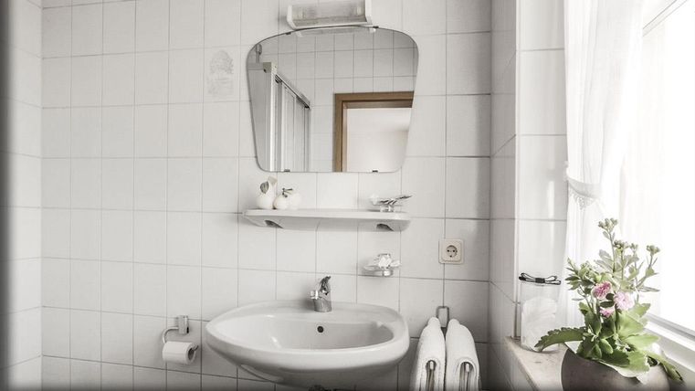 Une salle de bain lumineuse avec des carreaux blancs et un miroir rond au-dessus du lavabo. Sur l'étagère, des fleurs fraîches et des serviettes sont visibles.