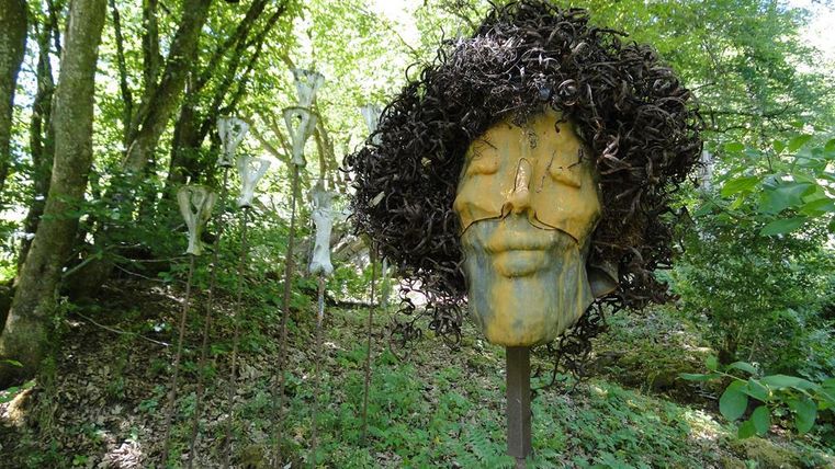 Eine faszinierende Skulptur mit einem menschlichen Gesicht und lockigem Haar steht im Wald. Im Hintergrund sind weitere kunstvolle Formen zwischen den Bäumen sichtbar.