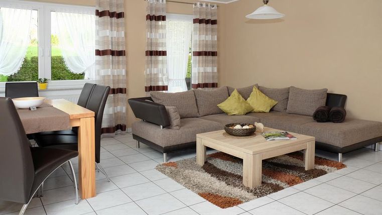 Ein modernes Wohnzimmer mit einer bequemen L-förmigen Couch und einem Holztisch. Große Fenster lassen viel Tageslicht herein und schaffen eine einladende Atmosphäre.