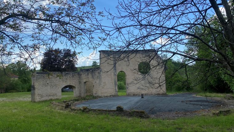 Eine alte Mauerruine umgeben von Bäumen und Wiese. Der Himmel ist blau mit einigen Wolken.