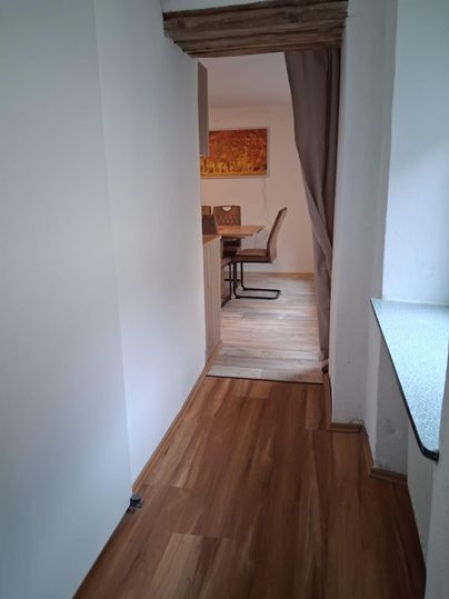 Un couloir lumineux avec un sol en bois, menant à un coin repas confortable. En arrière-plan, des chaises et une table sont visibles.