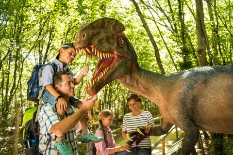 A family is standing in a forest and interacting with a dinosaur model. The children are excited while the adults are smiling and playing with the dinosaur.