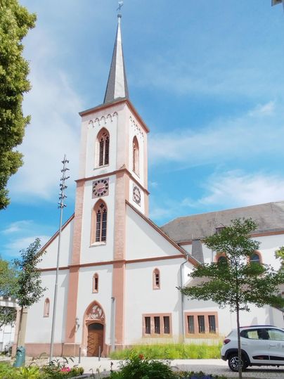 Eine schöne Kirche mit einem hohen Turm und einem klaren Himmel. Um die Kirche herum gibt es Bäume und einen Parkplatz.