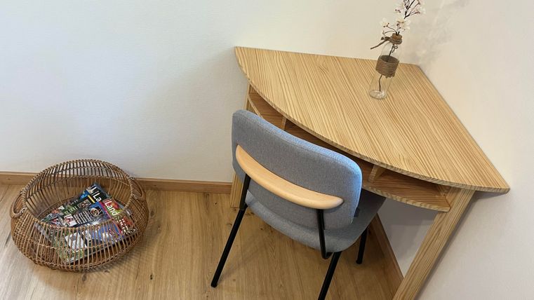 Un petit bureau en bois avec une chaise grise. À côté de la table, il y a un panier avec des magazines.