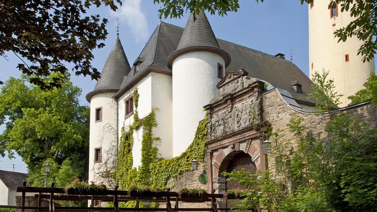 Eine malerische Burg mit Türmen und einem begrünten Eingang. Eine Holzbrücke führt über einen kleinen Gewässerlauf.
