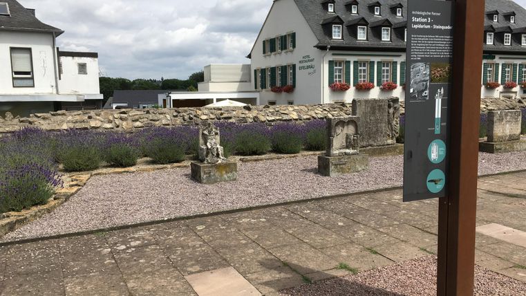 Eine ruhige Gartenlandschaft mit Lavendel und historischen Steinen. Im Hintergrund sind moderne und traditionelle Gebäude zu sehen.