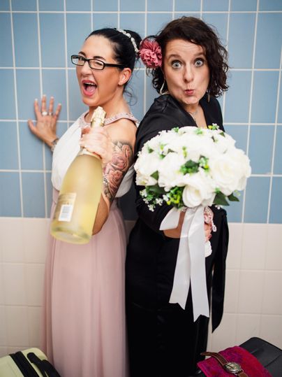 Zwei fröhliche Frauen posieren in einem Badezimmer. Eine hält eine Flasche Sekt und die andere einen Blumenstrauß.