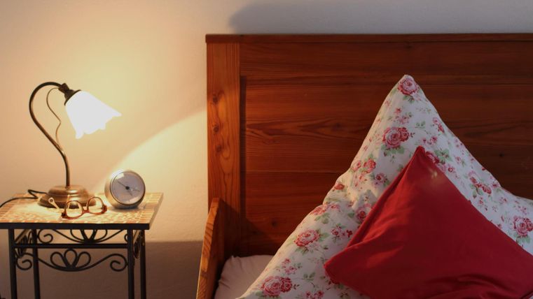 Ein gemütliches Bett mit floralem Bettzeug und einem roten Kissen. Neben dem Bett steht ein Tisch mit einer Lampe und einer Uhr.