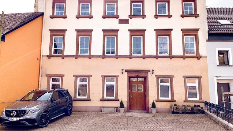 Un bâtiment de trois étages avec une façade claire et des cadres de fenêtres bruns. Devant la maison se trouve une voiture noire sur un parking pavé.