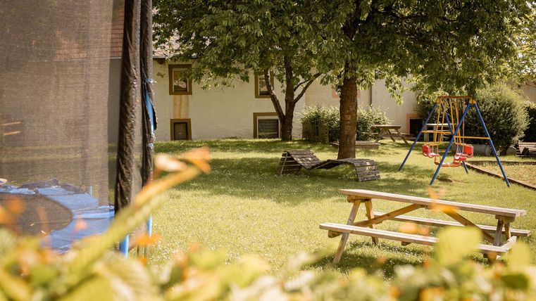 Een mooie tuin met een weide, een grote boom en een schommel. Aan de linkerkant staat een trampoline, en op de achtergrond zie je een huis.