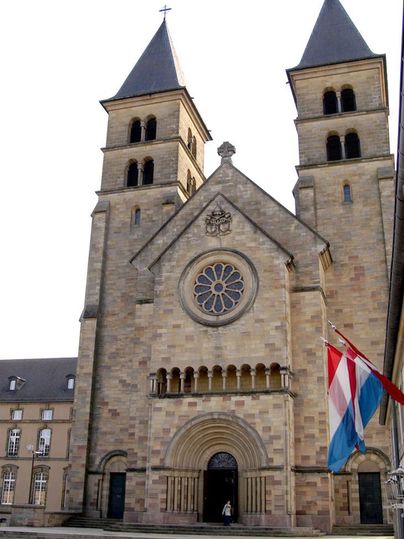 Une impressionnante église avec deux tours et une grande rosette. Devant l'église, les drapeaux du Luxembourg flottent.