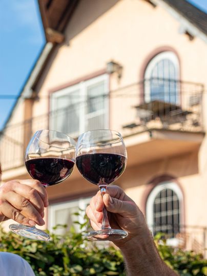 Zwei Gläser Rotwein werden anstoßen, während ein schönes Haus im Hintergrund zu sehen ist. Die Szene vermittelt eine festliche und entspannte Atmosphäre.
