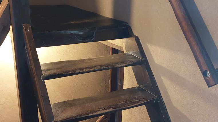 Een eenvoudige houten trap leidt naar een kleine ruimte. De muren zijn in een warme bruintint en de verlichting is zacht.