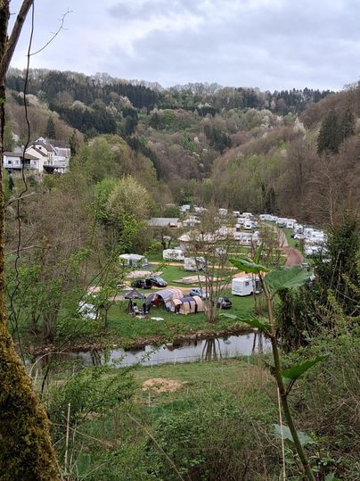 Un camping pittoresque dans une vallée verdoyante, entouré d'arbres. Des camping-cars sont alignés le long de la rivière, tandis que les collines en arrière-plan sont visibles.