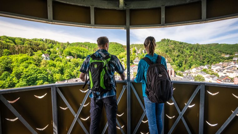 Zwei Personen mit Rucksäcken stehen auf einem Aussichtsturm und blicken auf eine grüne Landschaft und eine Stadt.