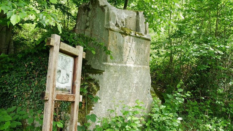 Ein altes Steinmonument im Wald, umgeben von grünen Blättern, mit einer Informationstafel daneben.