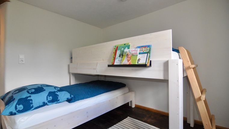 Ein gemütliches Hochbett in einem modernen Raum. Auf dem Bett liegt eine blaue Decke und ein Regal mit Büchern ist sichtbar.