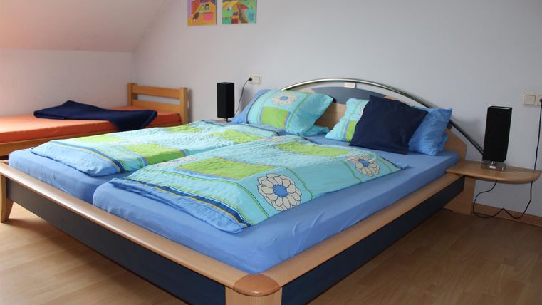 Een groot tweepersoonsbed met kleurrijke dekens en kussens. Op de achtergrond zijn een nachtkastje en een tweede bed te zien.