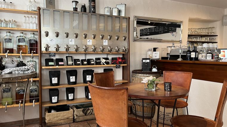 Un café accueillant avec une table en bois et des chaises. À l'arrière-plan, des étagères avec différentes boissons et accessoires sont visibles.