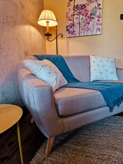 Een gezellige woonkamer met een lichte bank, waarop een blauwe deken en kussens liggen. Naast de bank staat een tafeltje en een lamp zorgt voor warm licht.