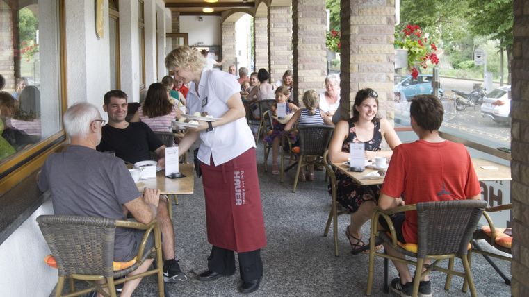 Une terrasse animée d'un restaurant avec des clients assis à des tables. Un serveur sert des plats et l'ambiance est conviviale et détendue.