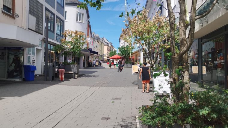 Eine belebte Einkaufsstraße mit Geschäften und Bäumen. Im Hintergrund sind Menschen und ein rotes Sonnenschirm zu sehen.