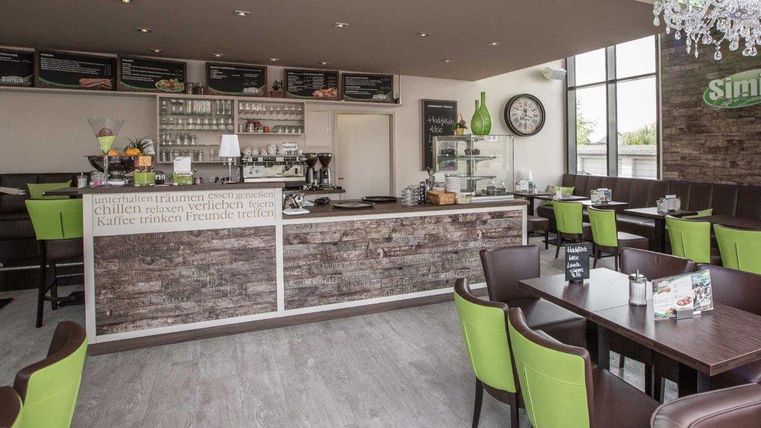 Un café moderne avec des tons de bois et de vert. Des tables et des chaises offrent une ambiance chaleureuse, tandis qu'un comptoir avec diverses boissons et snacks est visible en arrière-plan.