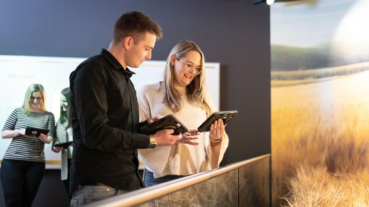 Zwei Personen stehen in einem Raum und schauen auf Tablets. Im Hintergrund ist eine dritte Person zu sehen, die ebenfalls ein Tablet benutzt.