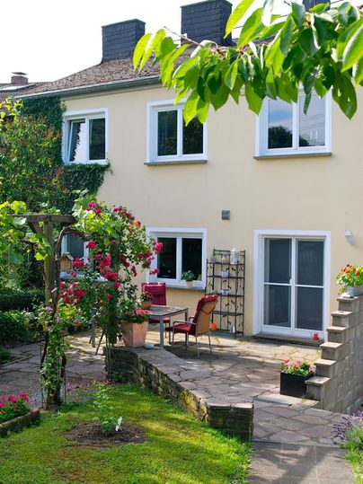 Ein gemütlicher Garten mit einer Terrasse und Blumenbüschen. Im Hintergrund steht ein helles Haus mit großen Fenstern.