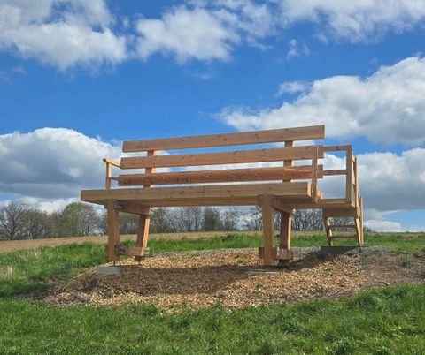 Een grote houten bank staat op een weide onder een blauwe lucht met wolken. De bank biedt een mooi uitzichtspunt op de omgeving.