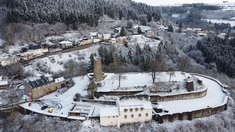 Eine schneebedeckte Burganlage umgeben von winterlicher Landschaft. Im Hintergrund sind die verschneiten Hügel und kleine Häuser sichtbar.