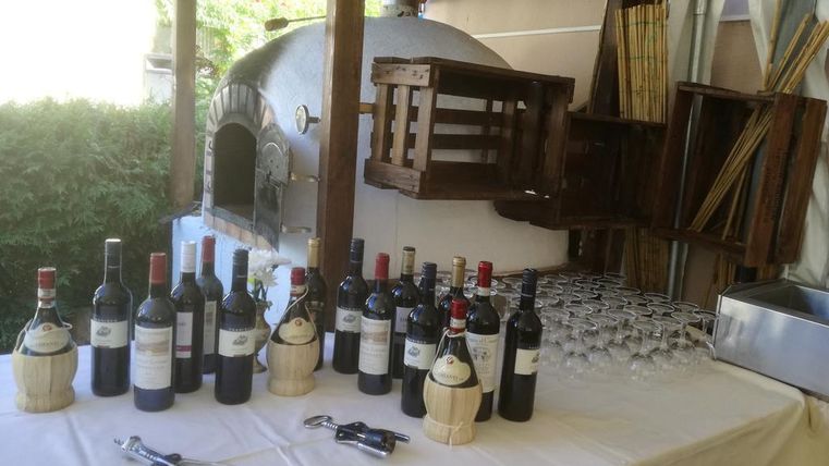 Une table joliment dressée avec différents vins rouges et des verres. En arrière-plan, il y a un four à pizza traditionnel.