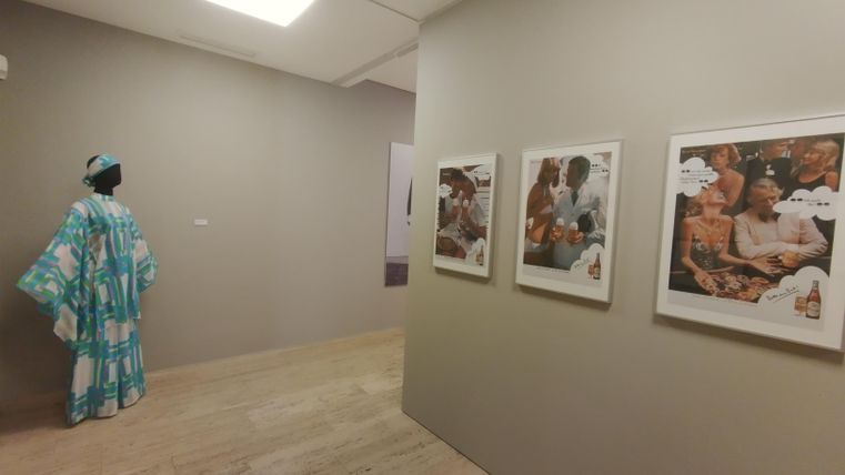 Ein Kunstraum mit grauen Wänden und einem Holzfußboden. An der Wand hängen mehrere gerahmte Fotos, während eine Figur in traditioneller Kleidung im Hintergrund steht.