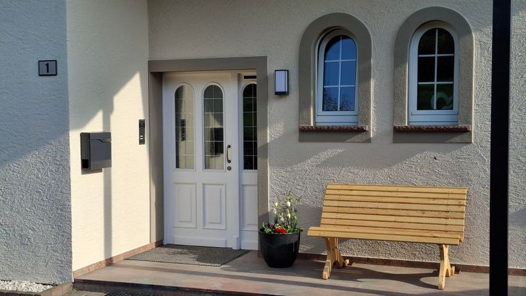 Eine einladende Haustür mit einem Holzbank und einem Blumentopf. Die Fassade hat runde Fenster und eine helle Farbgebung.